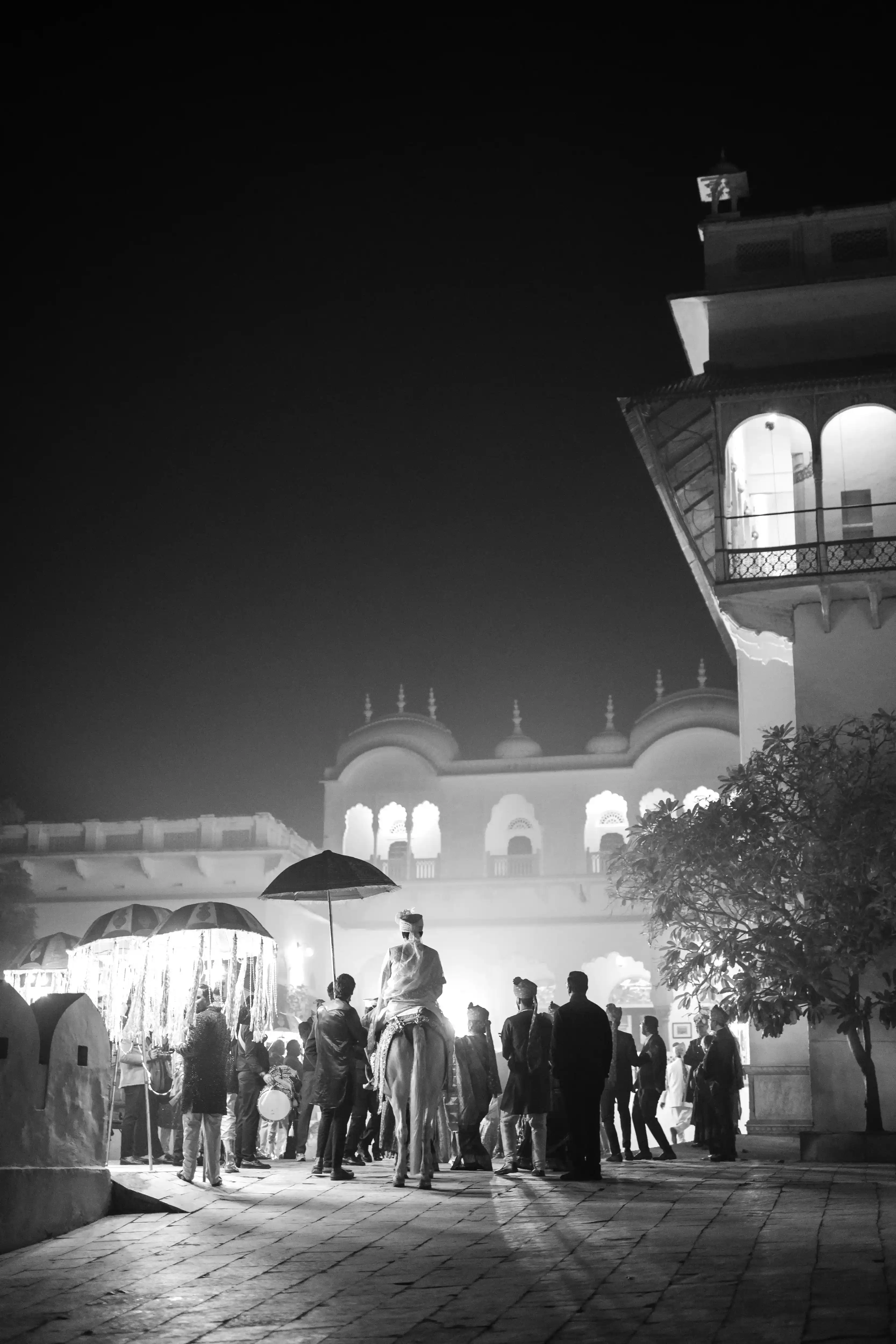 A timeless black and white photograph captures the scale of Mayank's royal baraat from behind, as the procession of guests follows the groom towards the entrance of Alsisar Mahal.
